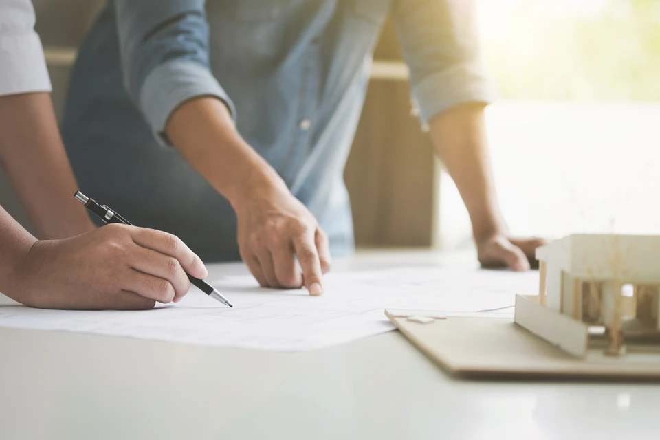 Sc&egrave;ne de travail collaboratif en int&eacute;rieur avec deux professionnels pench&eacute;s sur des plans architecturaux &eacute;tal&eacute;s sur une table