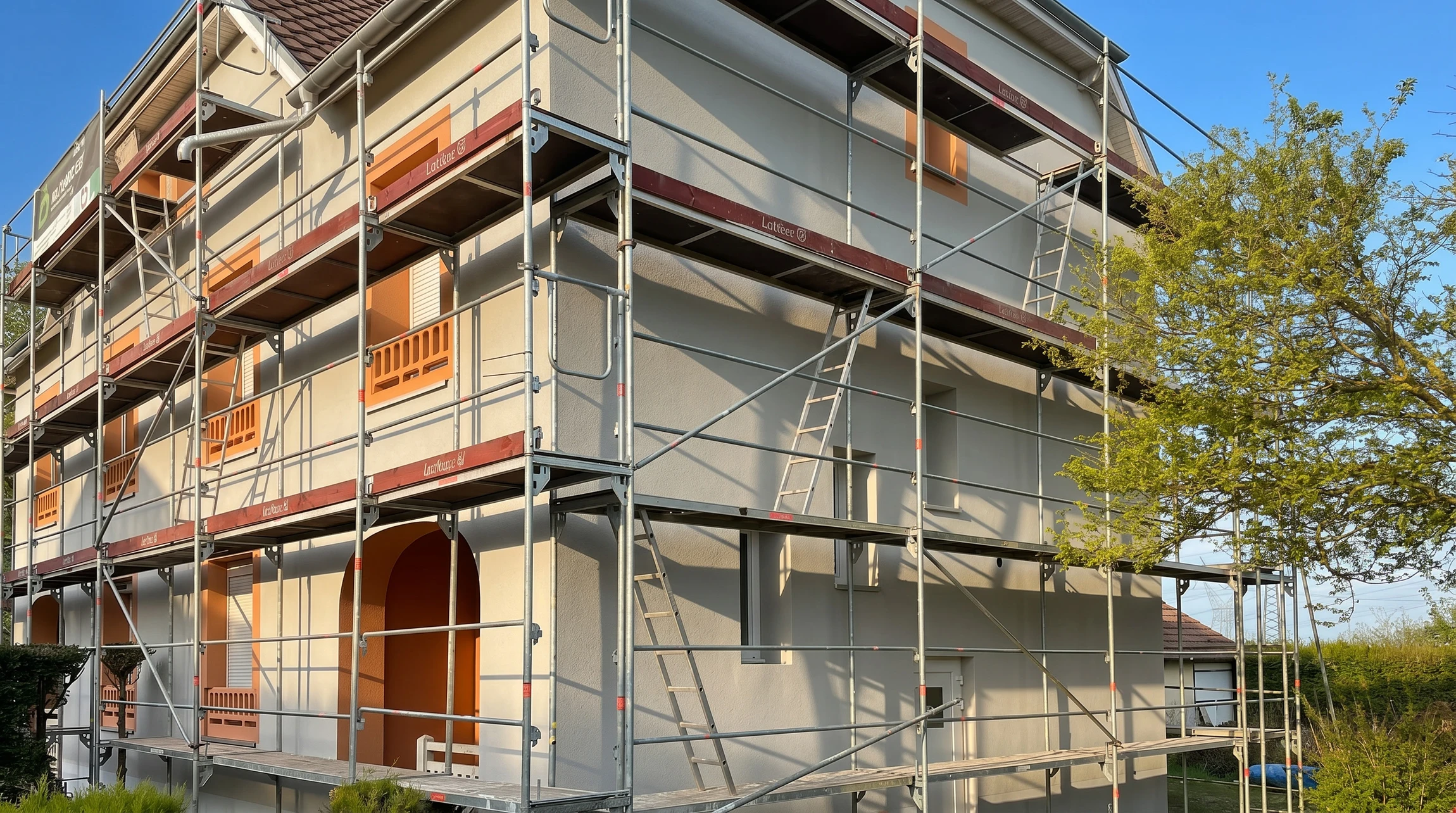 Scène de construction résidentielle avec ouvriers en casques sur chantier maison bois à ossature apparente, échafaudages métalliques, matériaux empilés au sol
